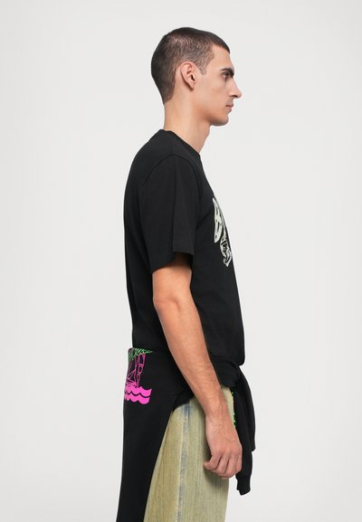 Schwarzes T-Shirt mit grafischem Design auf der Vorderseite, kombiniert mit hellen, strukturierten Hosen; auffälliges pinkes Logo-Detail auf der Rückseite.