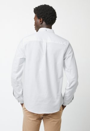 Homme aux cheveux courts et bouclés portant une chemise blanche à manches longues boutonnée et un pantalon beige, vu de dos.