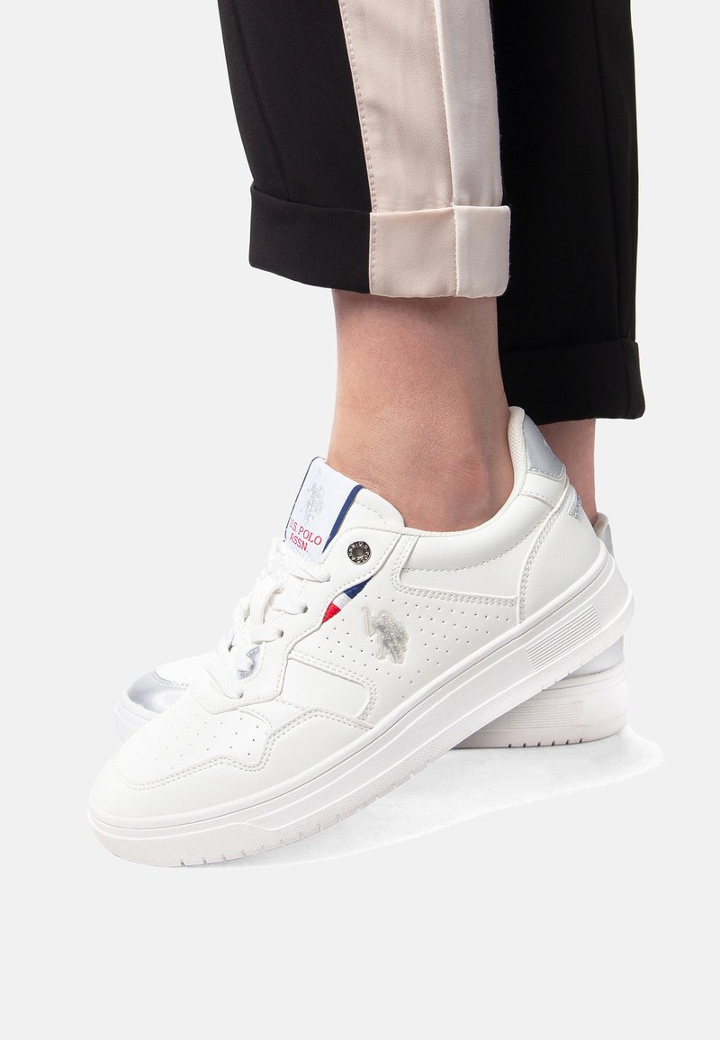 Hvide lavvandede sneakers med perforerede detaljer og logo, båret med sorte bukser med beige sidelister og op rullede manchetter.