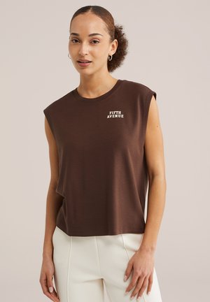 Jeune femme aux cheveux bouclés portant un haut marron sans manches marqué « Fifth Avenue » et un pantalon blanc, debout devant un fond beige uni.