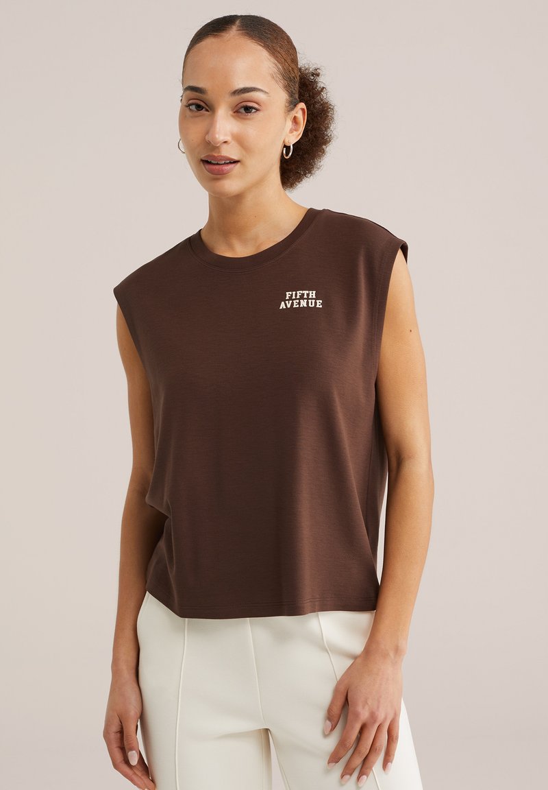 Fiatal nő göndör hajjal, barna ujjatlan felsőben, amelyen az "Ötödik sugárút" felirat olvasható, és fehér nadrágban, egyszínű bézs háttér előtt áll.