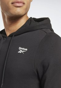 Sudadera negra de Reebok que presenta un logo blanco en el pecho, fabricada con un tejido suave y un acabado texturizado, y con una capucha de ajuste estándar.