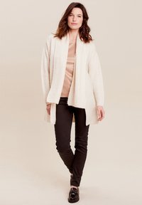 Licht beige, gemêleerde cardigan met open voorkant, lange mouwen en een ontspannen pasvorm, gecombineerd met een aangesloten roze top en zwarte broeken.