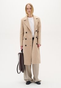 Bēša vilnas dubultā slāņa mēteli ar melnām pogām, sānu kabatām un platām atloks, kombinējot to ar gaiši bēšām biksēm un tumši brūnu somu.