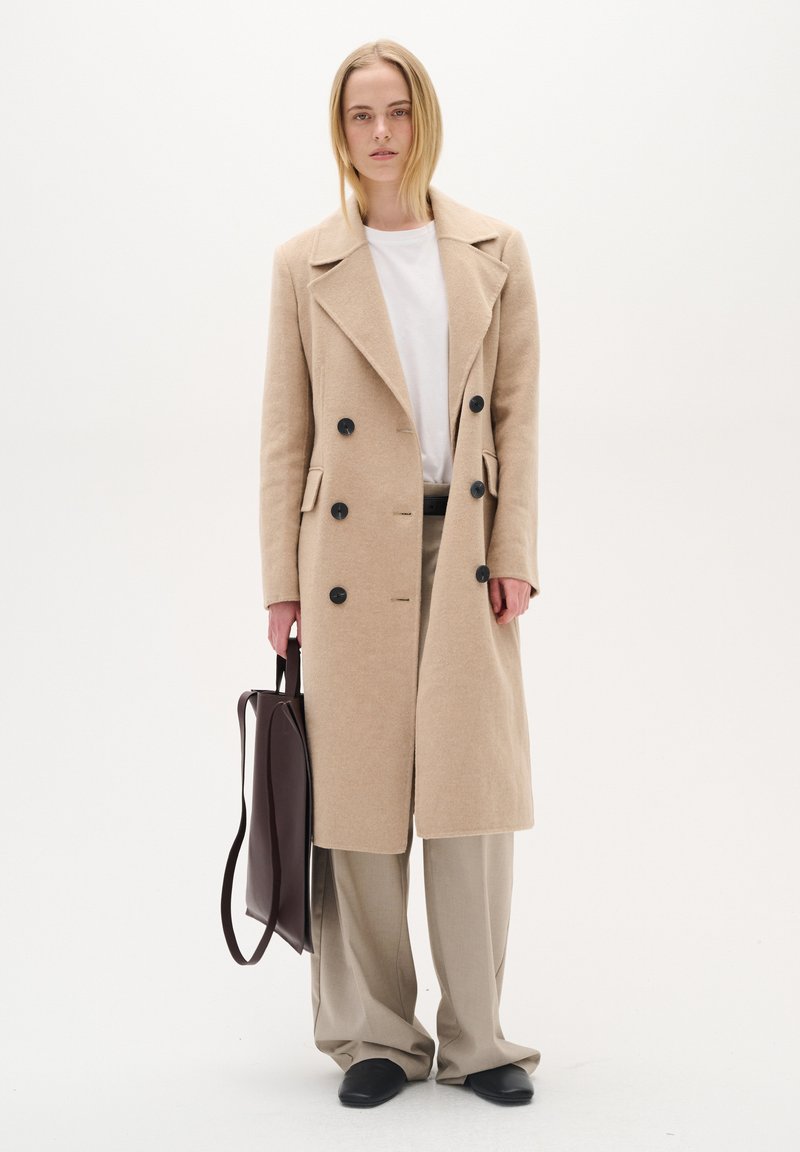 Bēša vilnas dubultā slāņa mēteli ar melnām pogām, sānu kabatām un platām atloks, kombinējot to ar gaiši bēšām biksēm un tumši brūnu somu.