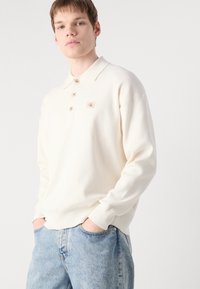 Κρεμώδης μακρυμάνικη πόλο μπλούζα με τρία κουμπιά και ribbed μανσέτες, διαθέτοντας ένα μικρό, τετράγωνο λογότυπο CK ραμμένο στο στήθος. Ορατά τζιν παντελόνια.