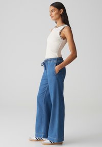 Pantalons en denim bleu à jambes larges avec une taille élastique et un cordon de serrage, associés à un haut blanc ajusté avec des bordures noires.
