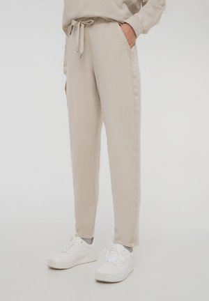 Pantaloni della tuta beige con cintura elastica e cordino, dotati di tasche laterali e strisce laterali a contrasto. Indossati con sneakers bianche.