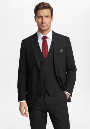 Man draagt een zwart driedelig pak met wit overhemd en rode stropdas, staand met één hand in de zak tegen een witte achtergrond.