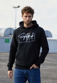 Schwarzer Hoodie mit Logo auf der Vorderseite, ausgestattet mit einer Kängurutasche und verstellbaren Kordeln. Kombiniert mit blauen Jeans aus strukturiertem Stoff.