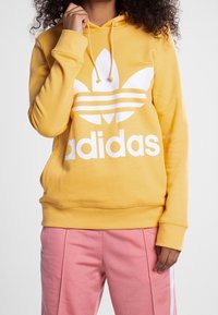 Person som bär en gul Adidas-hoodie med vit logotyp och rosa byxor med vita ränder, stående mot en enfärgad bakgrund.