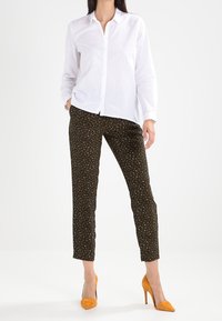 Vit långärmad skjorta med knappslå fram, tillsammans med mörkgrön mönstrad byxor och gula högklackade skor.