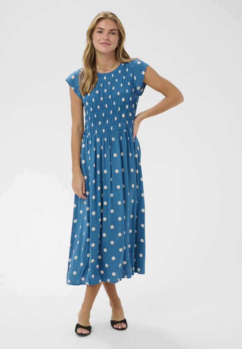 Femme portant une robe midi bleue à pois blancs, manches courtes et corsage smocké, debout avec la main sur la hanche et des talons noirs ouverts aux orteils.