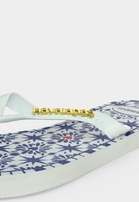 Λευκή σαγιονάρα με μπλε γεωμετρικά και floral σχέδια στη σόλα, λευκές υφασμάτινες λουράκια και χρυσό λογότυπο "havaianas" στο λουράκι.