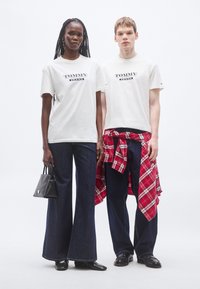 Λευκά βαμβακερά t-shirts με το λογότυπο "TOMMY JEANS", σε συνδυασμό με σκουρόχρωμο τζιν παντελόνι καμπάνα. Ένα μοντέλο κρατά μια μικρή μαύρη τσάντα· το άλλο έχει μια κόκκινη καρό πουκάμισο δεμένη στη μέση.