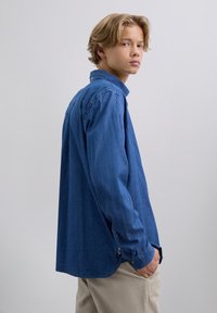 Joven con cabello rubio que lleva una camisa de mezclilla azul y pantalones beiges, de pie de lado con una mano en el bolsillo contra un fondo gris.