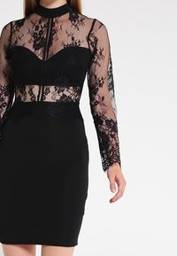 Svart klänning med en spetskorsett, långa ärmar, hög halsringning och en figursydd kjol. Spetsdetaljer framhäver midjan och ärmarna.