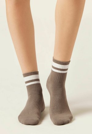 Nohy v hnědých kotníkových ponožkách s dvěma bílými pruhy u lemu, stojící na jemném neutrálním pozadí.