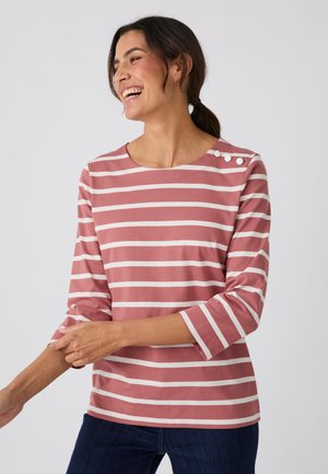Mujer sonriente con una camisa de rayas horizontales rosas y blancas, con mangas tres cuartos y botones en el hombro izquierdo.