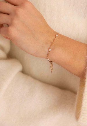 Handgelenk mit zartem Armband aus Goldkette mit kleinen weißen Perlen über einem cremefarbenen, gestrickten Ärmel.