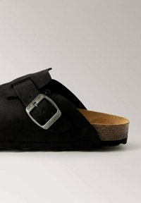 Black suede sandal featuring a cork footbed and a silver metal buckle. The design is open-toed with a textured surface and sturdy sole.