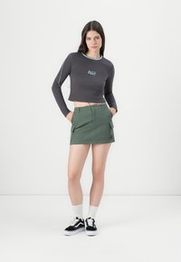 Korte lange mouwen top in donkergrijs met turquoise accenten; gecombineerd met een groene cargorok met zakken en een logo patch. Zwarte sneakers met witte zolen.