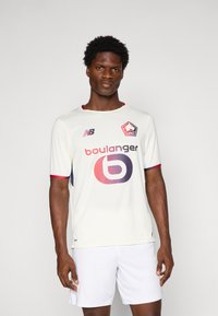 Vyrų modelis dėvi baltą LOSC Lille futbolo marškinėlį su raudonomis detalėmis, Boulanger logotipu ir baltais šortais, stovintis prieš vienodą pilką foną.
