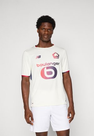 Mandlig model iført hvid trøje fra LOSC Lille med røde accenter, Boulanger-logo og hvide shorts, stående mod en ensfarvet grå baggrund.