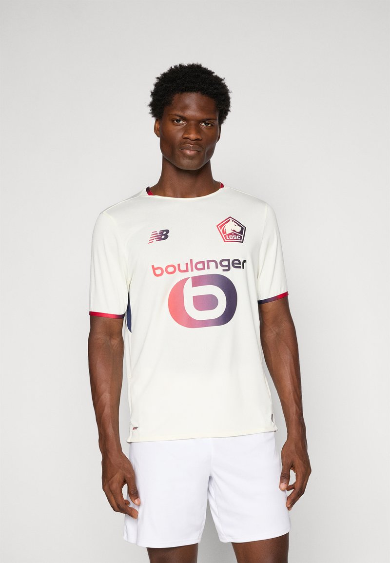 Vyrų modelis dėvi baltą LOSC Lille futbolo marškinėlį su raudonomis detalėmis, Boulanger logotipu ir baltais šortais, stovintis prieš vienodą pilką foną.