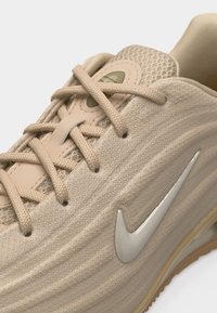 Κοντινή λήψη μπεζ αθλητικού παπουτσιού Nike που δείχνει την υφή του υφάσματος, τα κορδόνια, τη μαλακή γλώσσα και το ασημί λογότυπο Nike swoosh στο πλάι.