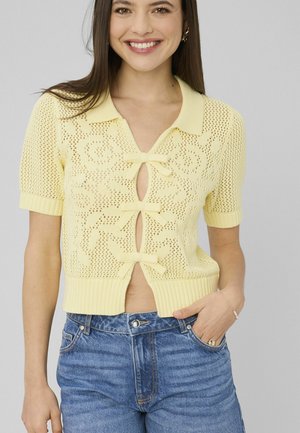 Jeune femme souriante, portant un haut tricoté jaune à manches courtes avec trois nœuds à l'avant et un jean bleu, main dans la poche, sur fond uni.