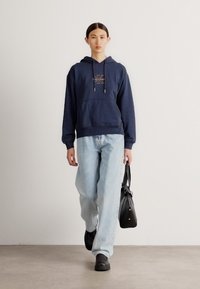 Sweatshirt capuz azul-marinho com bolso frontal, combinado com jeans retos azuis claros e calçado preto, a levar uma mala preta.
