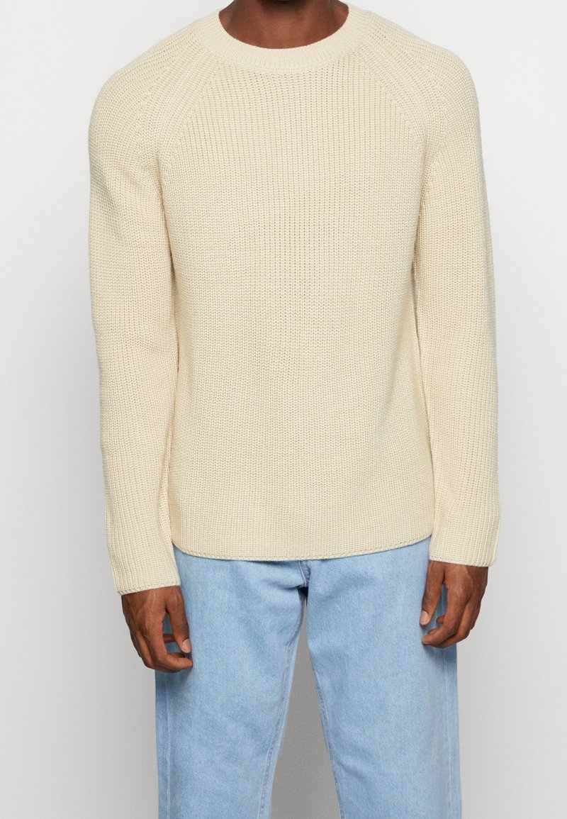 Homme portant un pull en maille côtelée couleur crème et un jean bleu clair, debout devant un fond blanc uni.