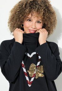 Sudadera negra con un alto cuello, que presenta un diseño de corazón y lazo hecho de lentejuelas en dorado, rojo y blanco. La tela parece suave y gruesa.