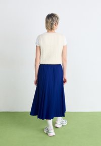 Kremfarget strikket vest over en hvit t-skjorte, kombinert med en knelengd plissert blå skjørt. Modellen har på seg grå og rosa joggesko med hvite sokker.