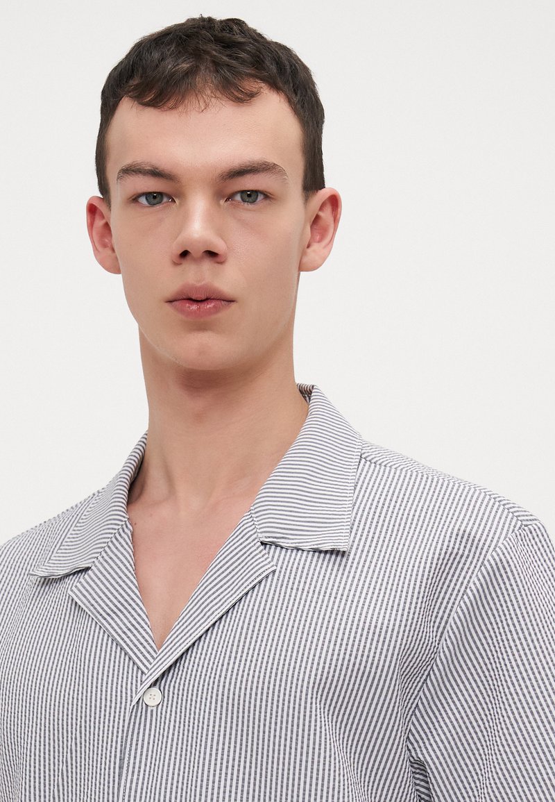 Chemise rayée en marine et blanc, dotée d'un col en V et d'un bouton unique. Tissu léger avec une texture lisse et des détails minimaux.
