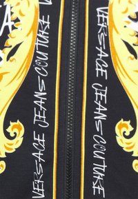 Svart tyg med djärva gula och vita mönster, inklusive en dekorativ kant och texten "Versace Jeans Couture." Mittendragkedjefunktion.