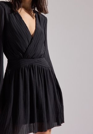 Femme portant une robe noire plissée à manches longues avec un décolleté en V profond et une taille cintrée sur un fond clair uni.