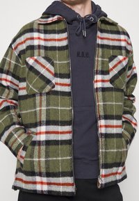 Zielona flanelowa kurtka w kratę, z pomarańczowymi, białymi i czarnymi akcentami. Posiada dwie kieszenie na piersi oraz zamek błyskawiczny. Nosi się na granatowym hoodie.