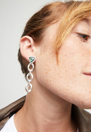 Pendientes de plata con una piedra triangular verde en la parte superior y cadenas ovaladas entrelazadas debajo, exhibiendo metal pulido y texturas suaves.