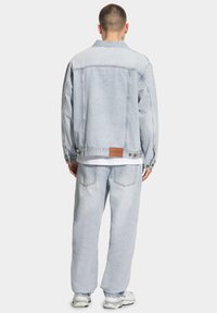 Jasnoniebieska dżinsowa kurtka o luźnym kroju, z widocznym szwem i metką marki. Dopasowana do jasnoniebieskich dżinsowych spodni i trampek.