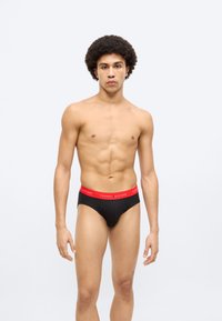 Μαύρα εσώρουχα με κόκκινη ζώνη μέσης που φέρει το λογότυπο "TOMMY HILFIGER", κατασκευασμένα από μαλακό ύφασμα, με εφαρμοστή σχεδίαση και χωρίς επιπλέον σχέδια ή διακοσμήσεις.