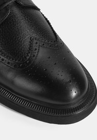 Schwarze Lederschuhe mit glatter Oberfläche, dekorativen Perforationen und strukturiertem Obermaterial. Sie verfügen über eine geriffelte Gummisohle und eine abgerundete Zehenpartie.