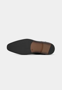 Semelle de chaussure présentant une semelle extérieure en caoutchouc noir texturé avec une zone d'accent lisse marron, une forme allongée et pointue, et une marque minimale.