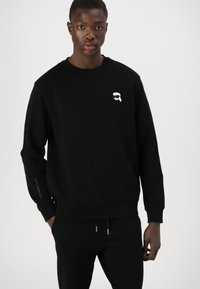 Črna kapuca z okroglim izrezom in dolgimi rokavi. Ima belo grafiko na prsih in besedilo vzdolž rokava. Mehka tekstura tkanine.