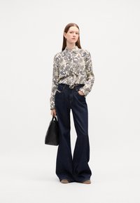 Long-sleeve paisley-patterned shirt in cream, blue, and gold, paired with dark blue flared denim pants. Black handbag with woven texture.