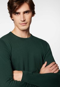 Zielony sweter o prążkowanej strukturze z klasycznym okrągłym dekoltem, długimi rękawami i teksturowanym materiałem, noszony przez osobę z założonymi rękami.