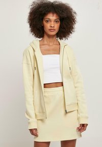 Sudadera amarilla con cremallera y un bolsillo frontal, combinada con una falda corta amarilla a juego. Top corto blanco debajo. Textura de algodón suave.