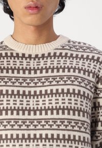 Stickad tröja med en krämfärgad bas och intrikat brunt geometriskt mönster. Ribbad rundad halsringning och mjuk textur. Inga synliga logotyper eller detaljer.