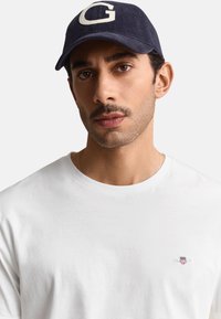 Mann mit kurzen dunklen Haaren und Schnurrbart, trägt ein weißes T-Shirt mit kleinem Logo und eine dunkelblaue Kappe mit großem "G" vorne.
